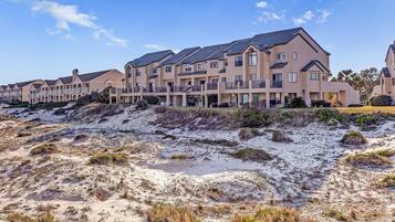 A row of modern multi-story buildings with balconies, situated on a sandy terrain with sparse vegetation.