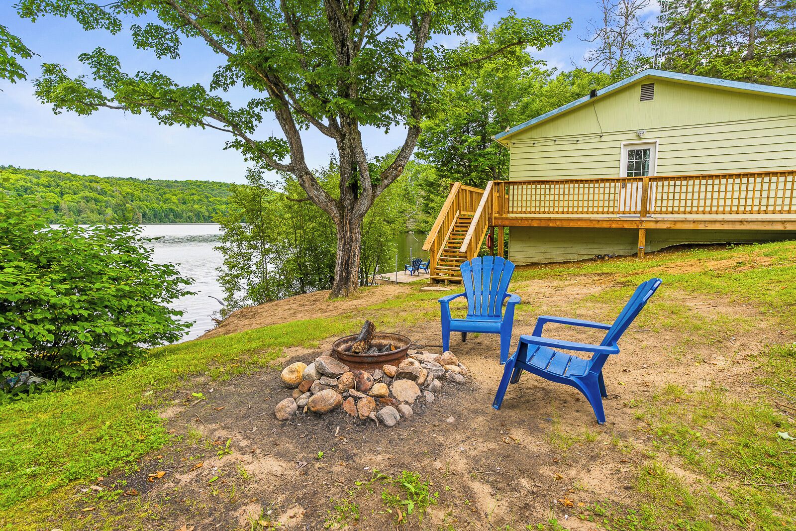 A lakeside cabin with a deck, a fire pit, and two Adirondack chairs.