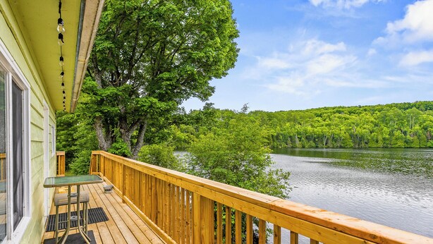 A wooden deck with a glass table and chairs, overlooking a lake surrounded by trees.