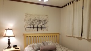 Ein Schlafzimmer mit einem hölzernen Bett, einem Nachttisch mit Lampe, einer Wandkunst und einem Fenster mit Vorhängen.