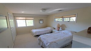 Un dormitorio con dos camas individuales, un escritorio y una silla. Hay ventanas con persianas, una puerta y una lámpara fijada a la pared.