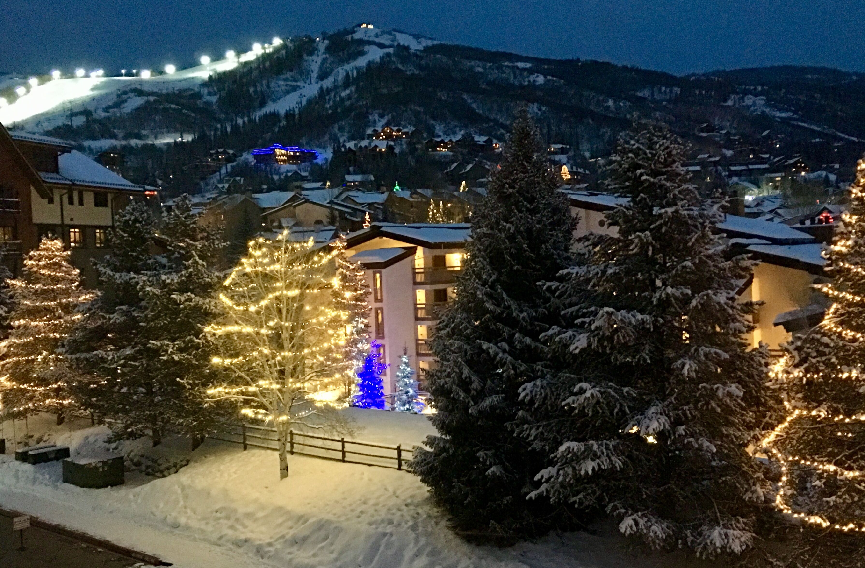 A snow-covered village with illuminated trees and buildings, a mountain in the background, and a ski lift.