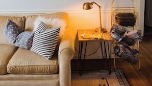 A living room with a beige sofa, patterned cushions, a small table with a lamp, a framed poster, and a wicker chair.