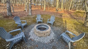 Een ronde vuurkuil omringd door vier Adirondack-stoelen in een bebost gebied.