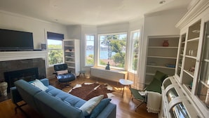 Een ruime woonkamer met een blauwe bank, een lederen fauteuil, een open haard en grote ramen met uitzicht op de omgeving.