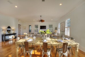 A dining room with a large table set for six, surrounded by chairs, with a ceiling fan above and a television on the wall.