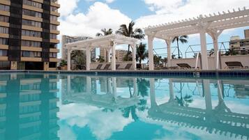 A swimming pool with a white pergola and lounge chairs, surrounded by tall buildings and palm trees.