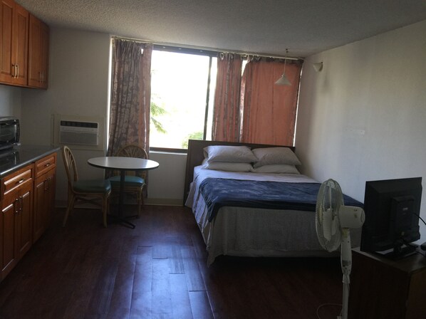 Una habitación compacta con cama, escritorio, silla, ventilador y televisión.