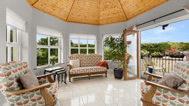A sunroom with a wooden ceiling, large windows, a sofa, two armchairs, and a potted plant.