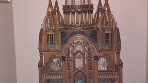 A wooden model of a building with multiple towers and a central dome, placed on a stand in a room with a window in the background.