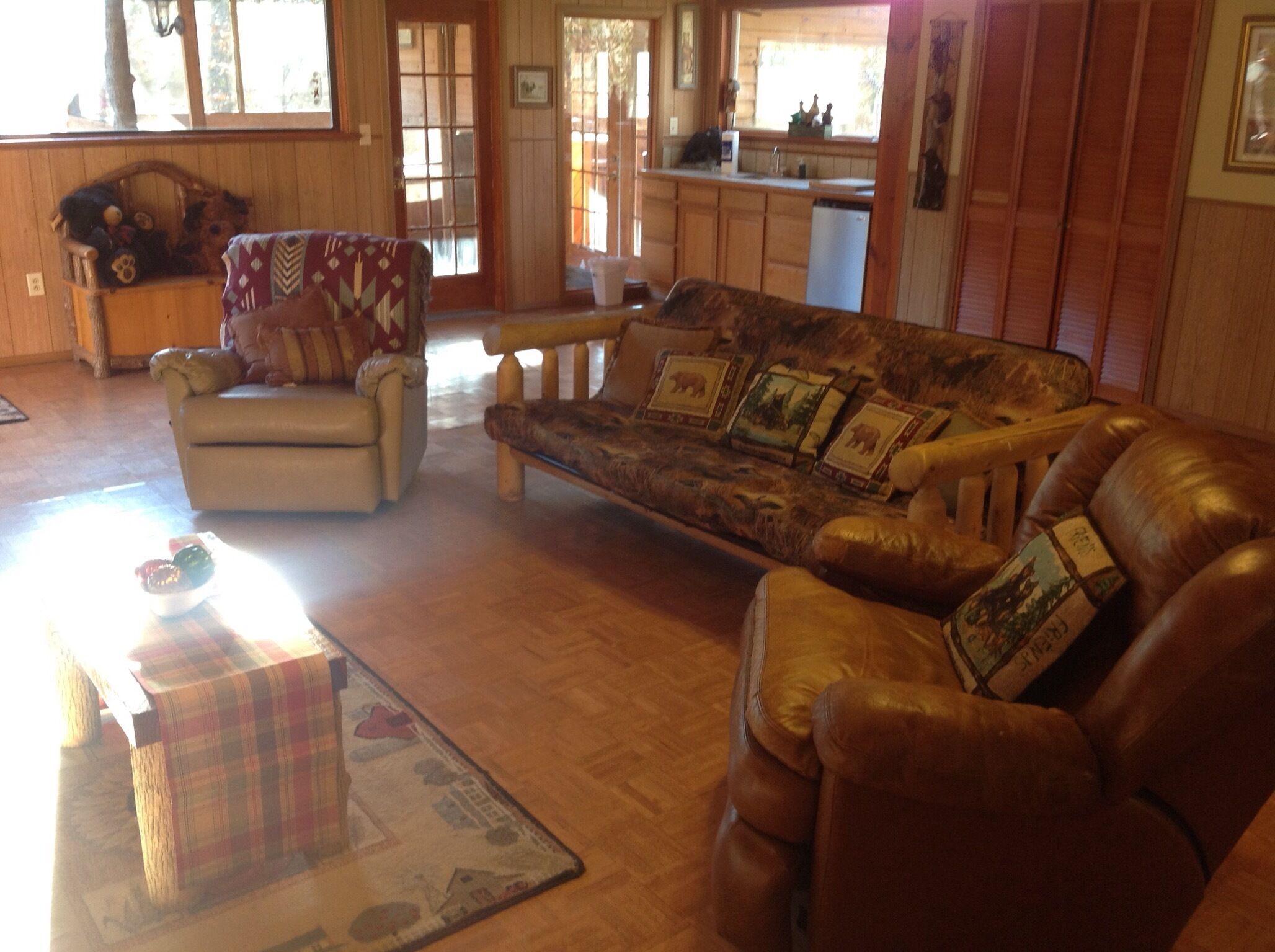 A cozy living room with a sofa, armchair, and a coffee table. There is a wooden dining table and a kitchen area in the background.