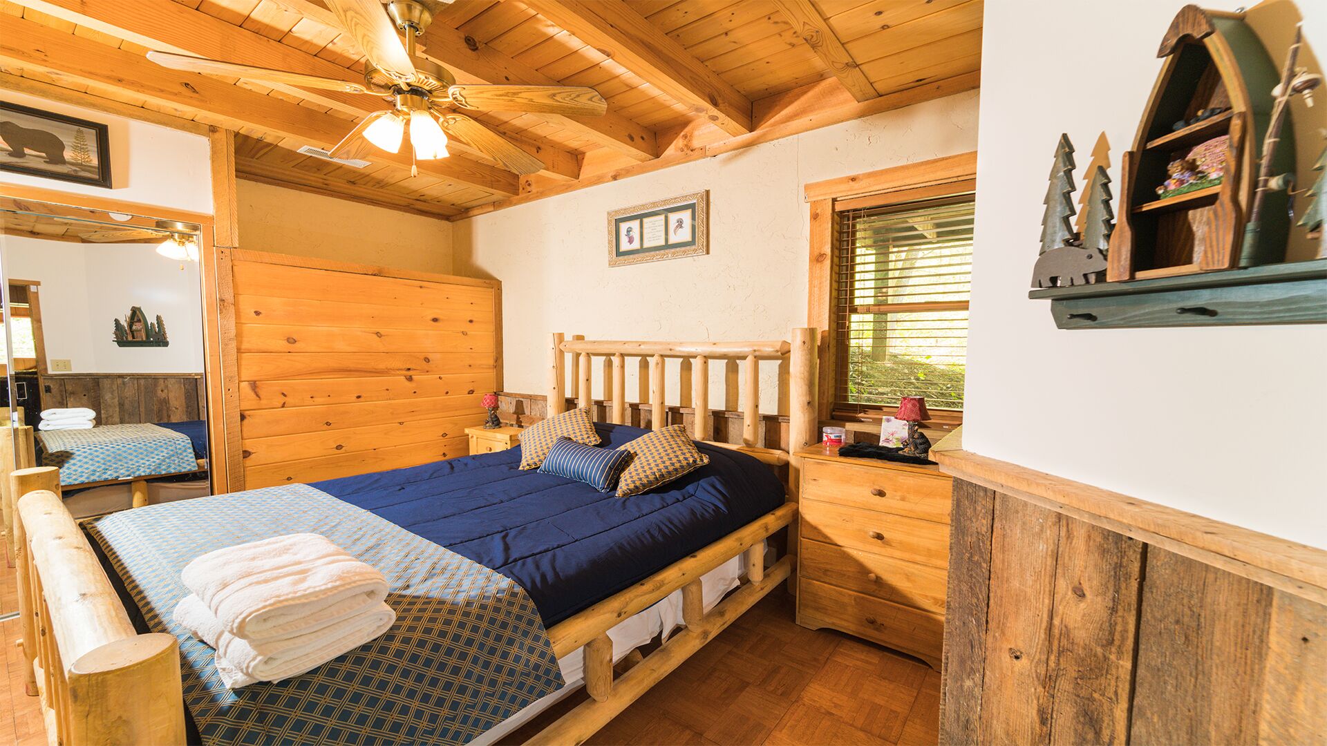 Une chambre dans une cabane en bois, comprenant un lit, une table de chevet, une commode, un ventilateur de plafond et une fenêtre avec des stores.