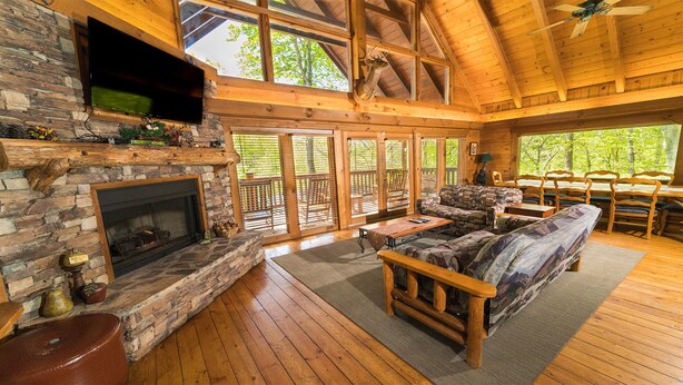 Un intérieur de cabane spacieux, agrémenté d’une cheminée en pierre, de meubles en bois et de grandes fenêtres offrant une vue sur le paysage extérieur.