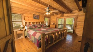 Une chambre dans une cabane en bois, comprenant un lit, une table de chevet, une commode, un ventilateur de plafond et une fenêtre avec des stores.