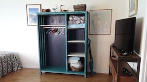 A teal wardrobe with hanging space and shelves, a television on a wooden stand, and a wicker basket on the top shelf.