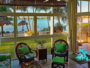 Relax in a serene room with a view of a calm body of water and palm trees, featuring a comfortable seating area with wicker chairs and a coffee table.