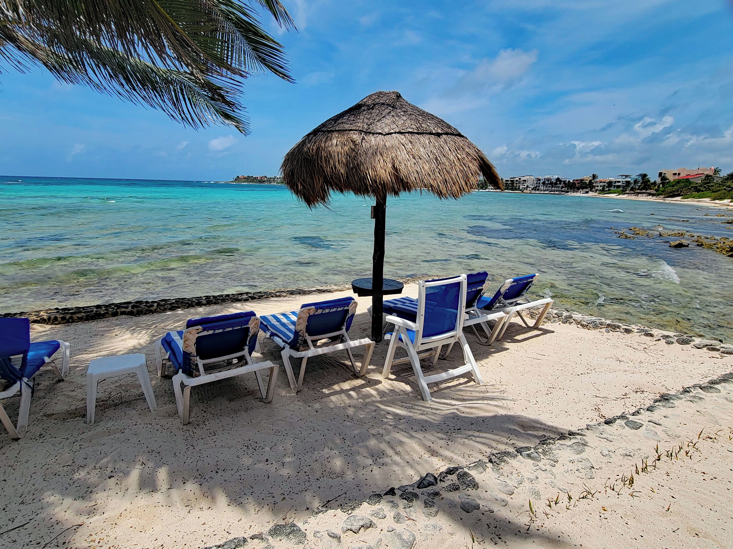 Een strand met helder turquoise water, een rieten parasol en blauwe ligstoelen.