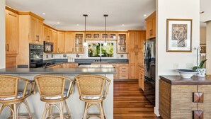 A modern kitchen with wooden cabinets, a central island with bar stools, and a large window providing a view of greenery.