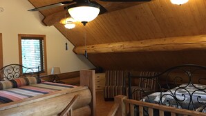 Un dormitorio de cabaña de madera con ventilador de techo, dos lámparas de noche, una cama con manta a rayas, una silla y una ventana con persianas.