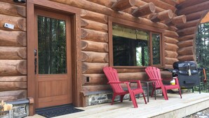 Una cabaña de troncos con puerta de madera, dos ventanas y un porche con sillas Adirondack rojas.