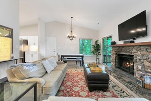 A living room with a stone fireplace, a flat-screen TV mounted above it, a sofa, a coffee table, and a dining area with a table and chairs.