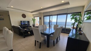 A modern living room with a dining area, a TV, and a large window overlooking the sea. - Stunning Oceanfront Pearl Tower Condo - Pool, Gym &  Sunsets - 3 beds/4baths (Mazatlán)