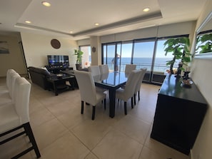 A modern living room with a dining area, a TV, and a large window overlooking the sea.