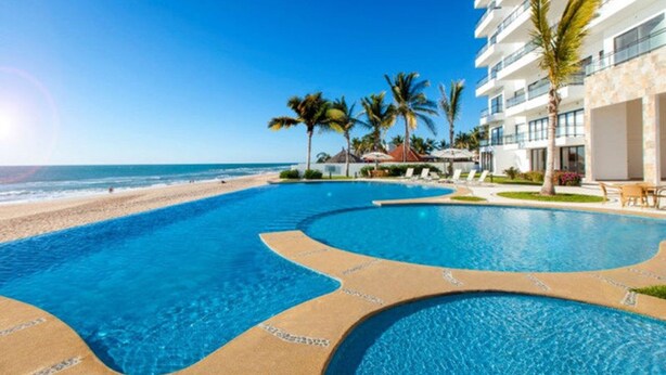 A swimming pool with a beach view, palm trees, and a modern building with multiple balconies. - Stunning Oceanfront Pearl Tower Condo - Pool, Gym &  Sunsets - 3 beds/4baths (Mazatlán)