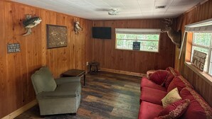 A room with wooden walls, a ceiling fan, a red sofa, a green armchair, and a wooden floor. - Camp House on Red Creek - Uncle Ralph’s Cabin Rentals - PRIVATE BEACH (Perkinston)