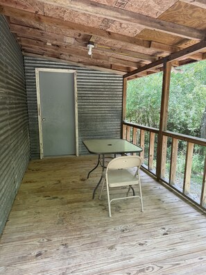 A covered outdoor area with a wooden table and a chair, a door, and a view of greenery through large windows.