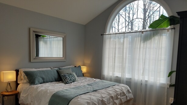 Un dormitorio con una cama grande, dos lámparas de noche, un espejo, una ventana con cortinas translúcidas y vistas a árboles.