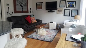Una sala de estar moderna con un sofá de cuero negro, una mesa de centro de madera y un otomano blanco y mullido. Hay un televisor montado en la pared, una mesita auxiliar y una alfombra estampada en el suelo.