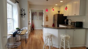 Una cocina moderna con suelo de madera, armarios blancos, una isla central con taburetes, luces colgantes y un refrigerador.