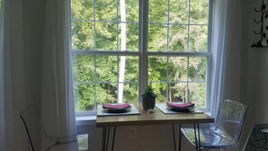 Una habitación con un ventanal grande, una mesa de madera, dos sillas transparentes y una planta en maceta.