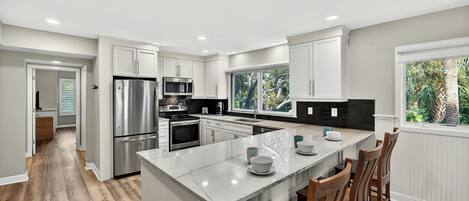 Una cocina moderna con armarios blancos, electrodomésticos de acero inoxidable, una isla grande con asientos y vistas a la vegetación a través de la ventana.