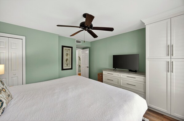 Un dormitorio con cama, ventilador de techo, televisor, armario y una puerta que da a otra habitación.