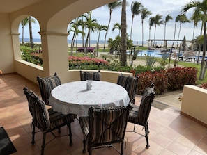 Une terrasse couverte avec une table ronde et des chaises à rayures, donnant sur une piscine et une plage bordées de palmiers, avec vue sur l’océan.