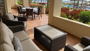 Un espacio de estar al aire libre techado con muebles de mimbre, una mesa con sillas y vistas a la playa y la piscina.