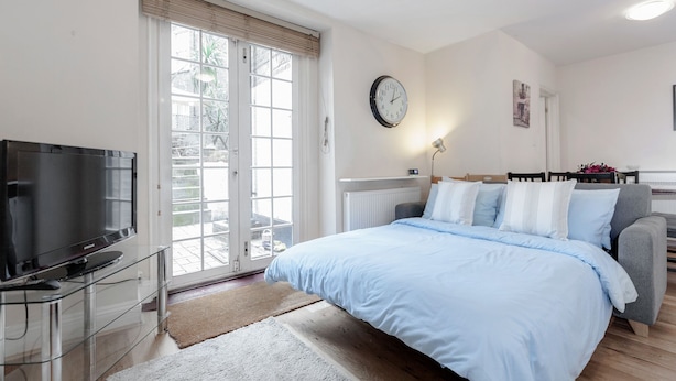 Une chambre à coucher avec un lit, un téléviseur posé sur un meuble en verre, une fenêtre avec des stores, un radiateur et une horloge au mur.
