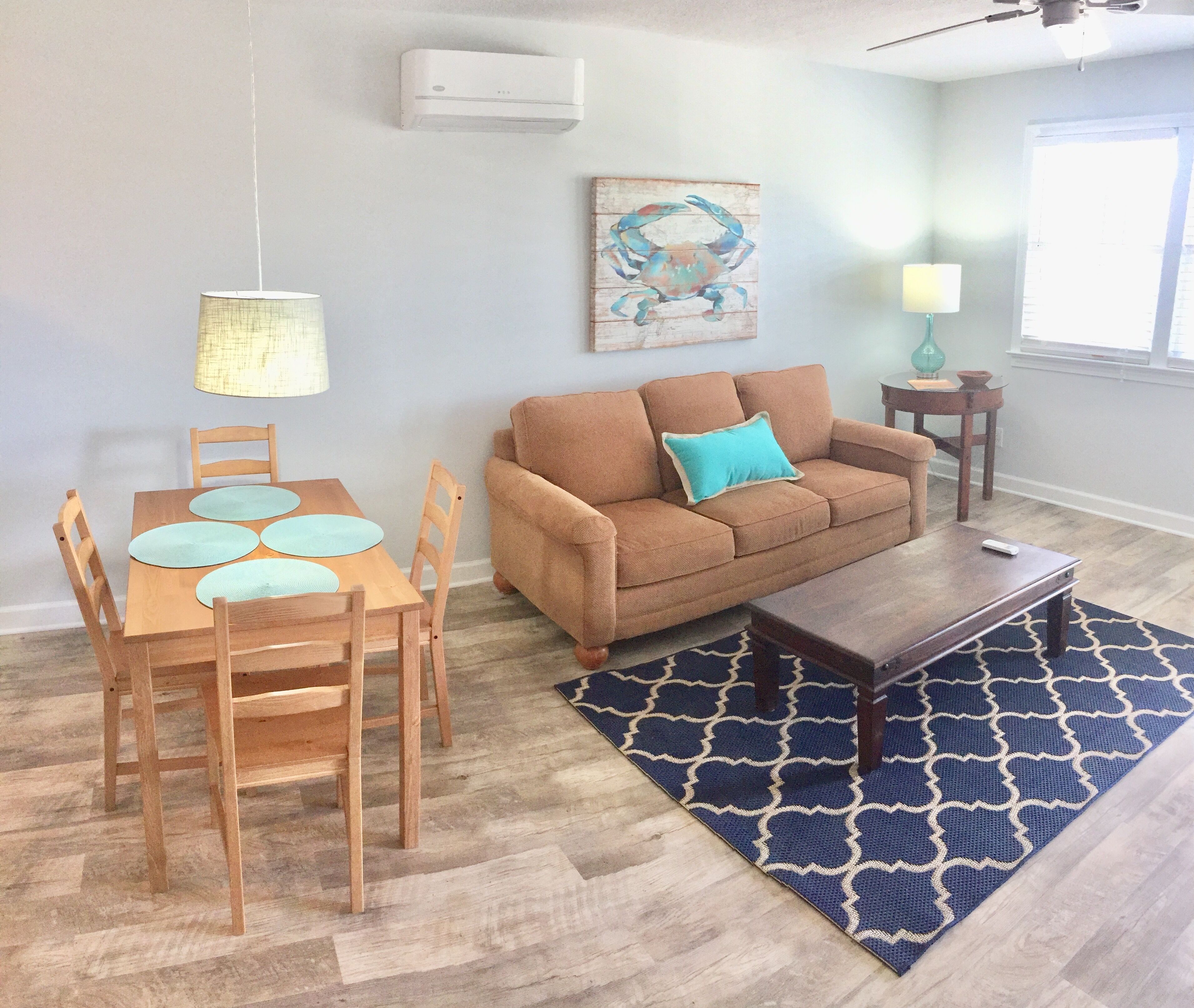 Una sala de estar con un sofá marrón, una mesa de comedor de madera con cuatro sillas, un tapete azul y un ventilador de techo.