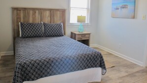 A bedroom with a bed, a nightstand, a lamp, and a painting on the wall. - Atlantic Oasis, less than 1 block to the beach! Private & Spacious, Full Kitchen (Nags Head)