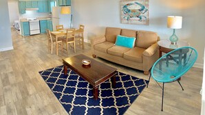 A living room with a sofa, coffee table, and a blue patterned rug. There is a dining area with wooden chairs and a table. A ceiling fan is visible, and a painting hangs on the wall. - Atlantic Oasis, less than 1 block to the beach! Private & Spacious, Full Kitchen (Nags Head)