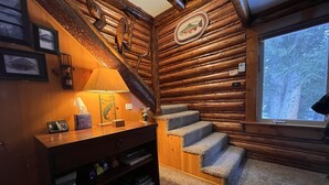 Interior de una cabaña de troncos con escalera de madera, un escritorio con lámpara y una ventana con vistas a árboles.