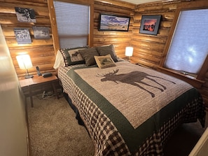 A cozy log cabin bedroom with a bed featuring a moose quilt, two bedside lamps, a nightstand, and framed pictures on the walls.