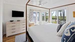 A bedroom with a large bed, a flat-screen TV, a wooden dresser, and a view of palm trees through the windows. - IDEAL Location at Windswept, 3BD Villa, Walk-Out to Beach! (Kiawah Island)