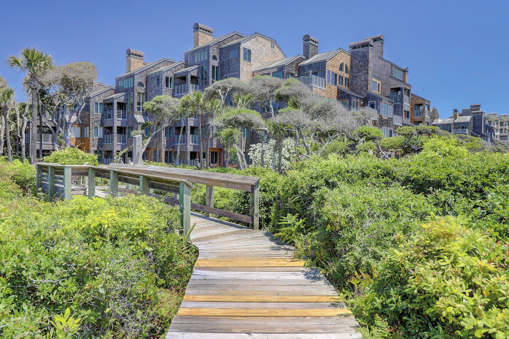 A wooden boardwalk leading to a multi-story building with balconies and a clear blue sky.