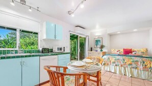 A modern kitchen with a dining area, a bed with a floral bedspread, and a view of greenery outside. - Haleakala Cottage - private hot tub, pool access (Haiku)