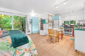 Een kamer met een bed, een eettafel met stoelen, een keuken met een fornuis en oven, en een groot raam met uitzicht op groen.