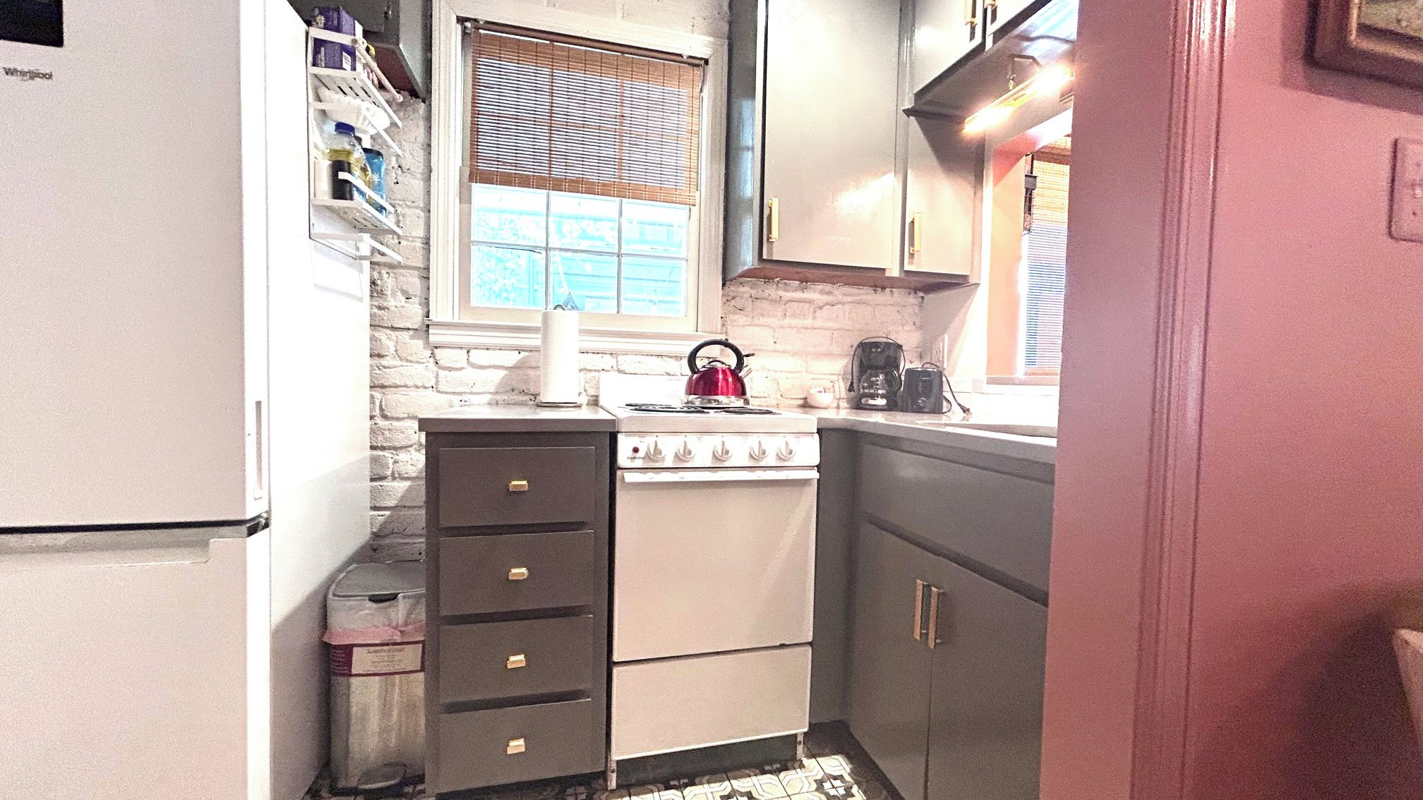 A compact kitchen with a white refrigerator, a stove, and a sink. There is a window with blinds, and a shelf with various items.