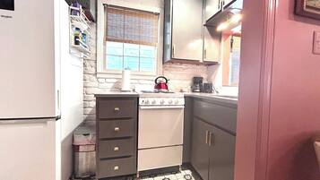 A compact kitchen with a white refrigerator, a stove, and a sink. There is a window with blinds, and a shelf with various items.